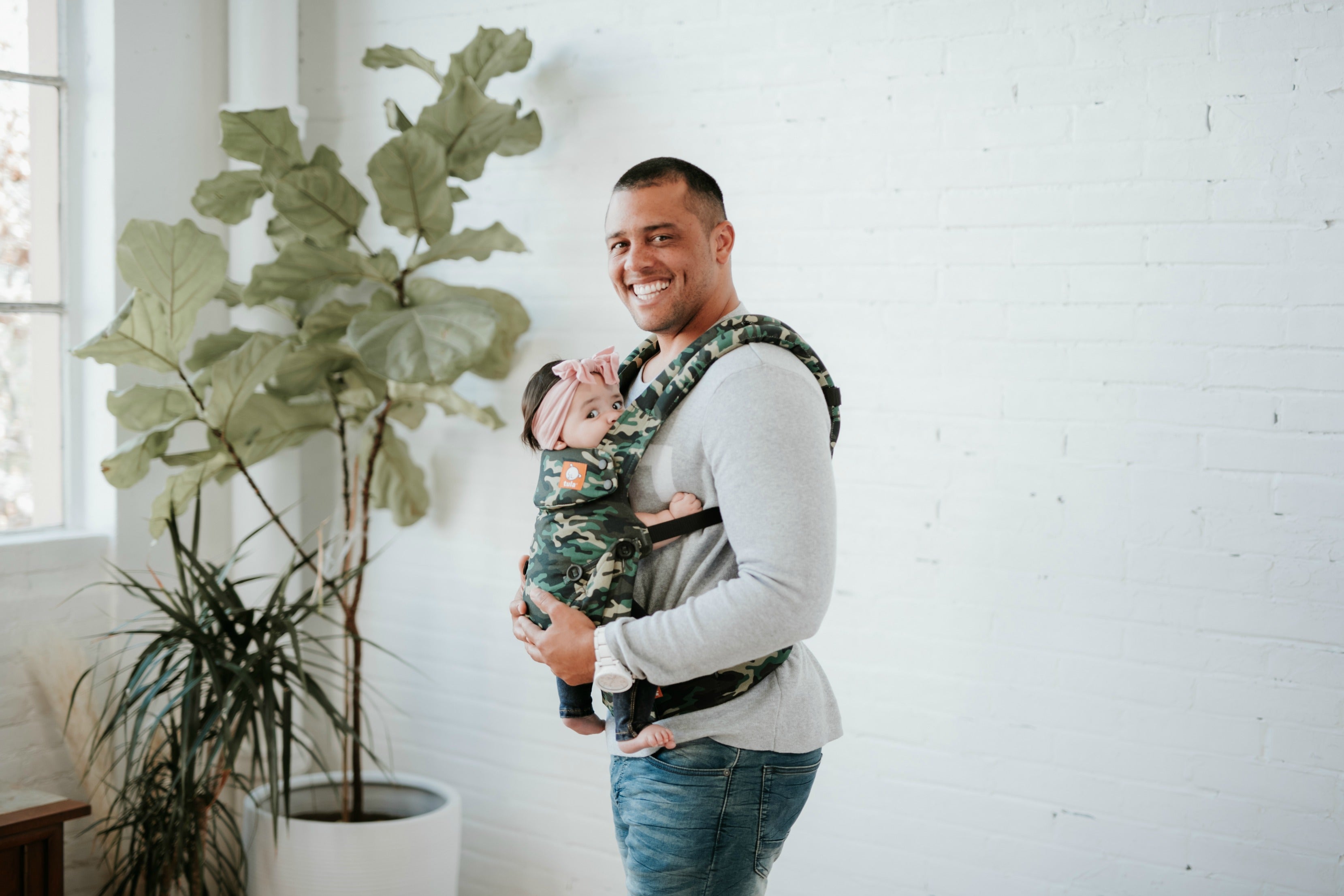 Father smiling while holding his baby in a camouflage-patterned soft structured carrier in front of a white brick wall.