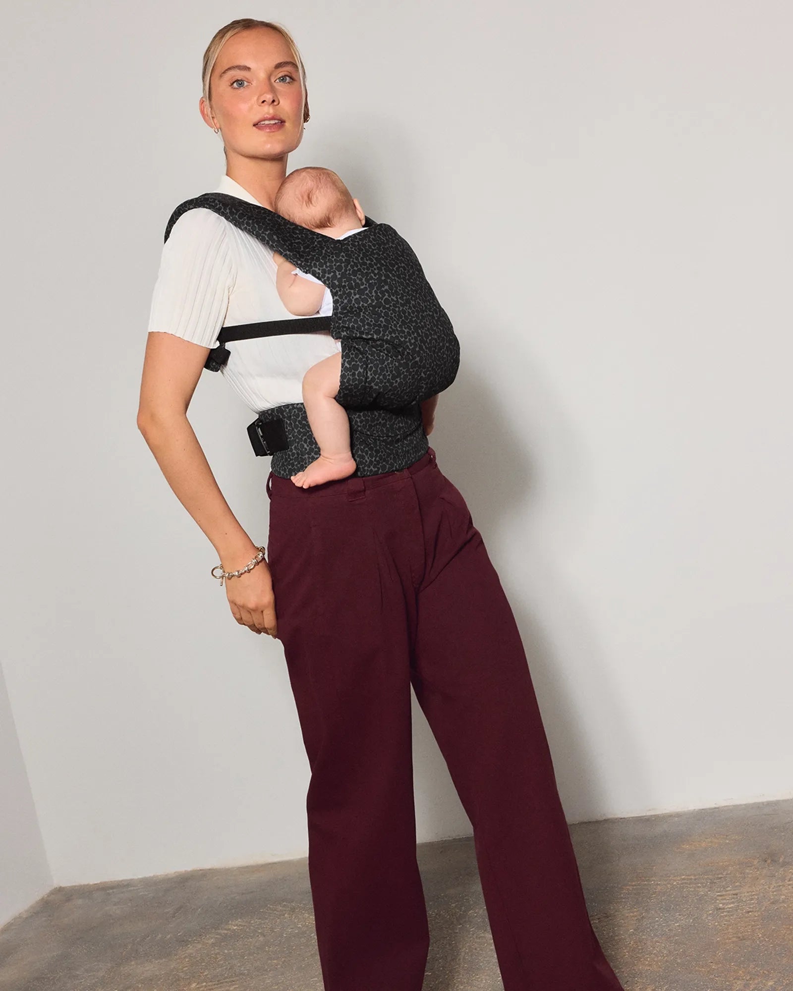 A woman wearing a dark gray patterned explore baby carrier with her newborn baby inside, styled with a white t-shirt and burgundy wide-leg trousers, standing against a neutral studio wall.