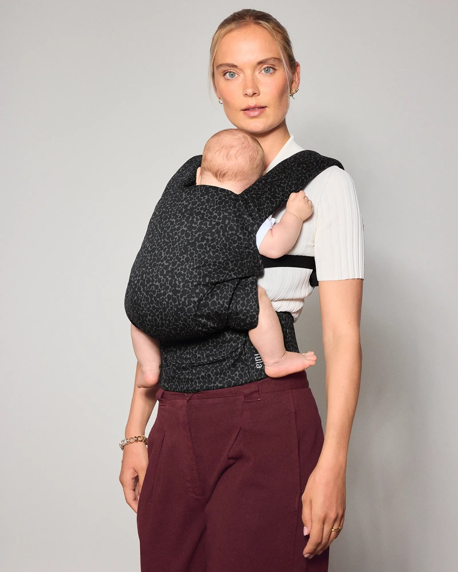 A woman wearing a black and gray speckled explore baby carrier carrying her newborn baby, standing against a plain light gray wall.