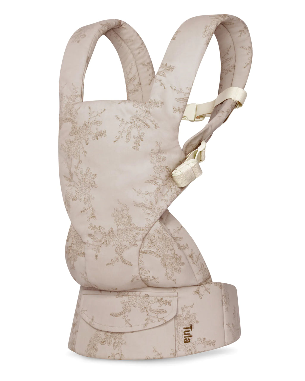 A Tula mesh baby carrier in a light beige fabric with a delicate floral toile pattern, featuring cream-colored buckle details and a matching front pouch.
