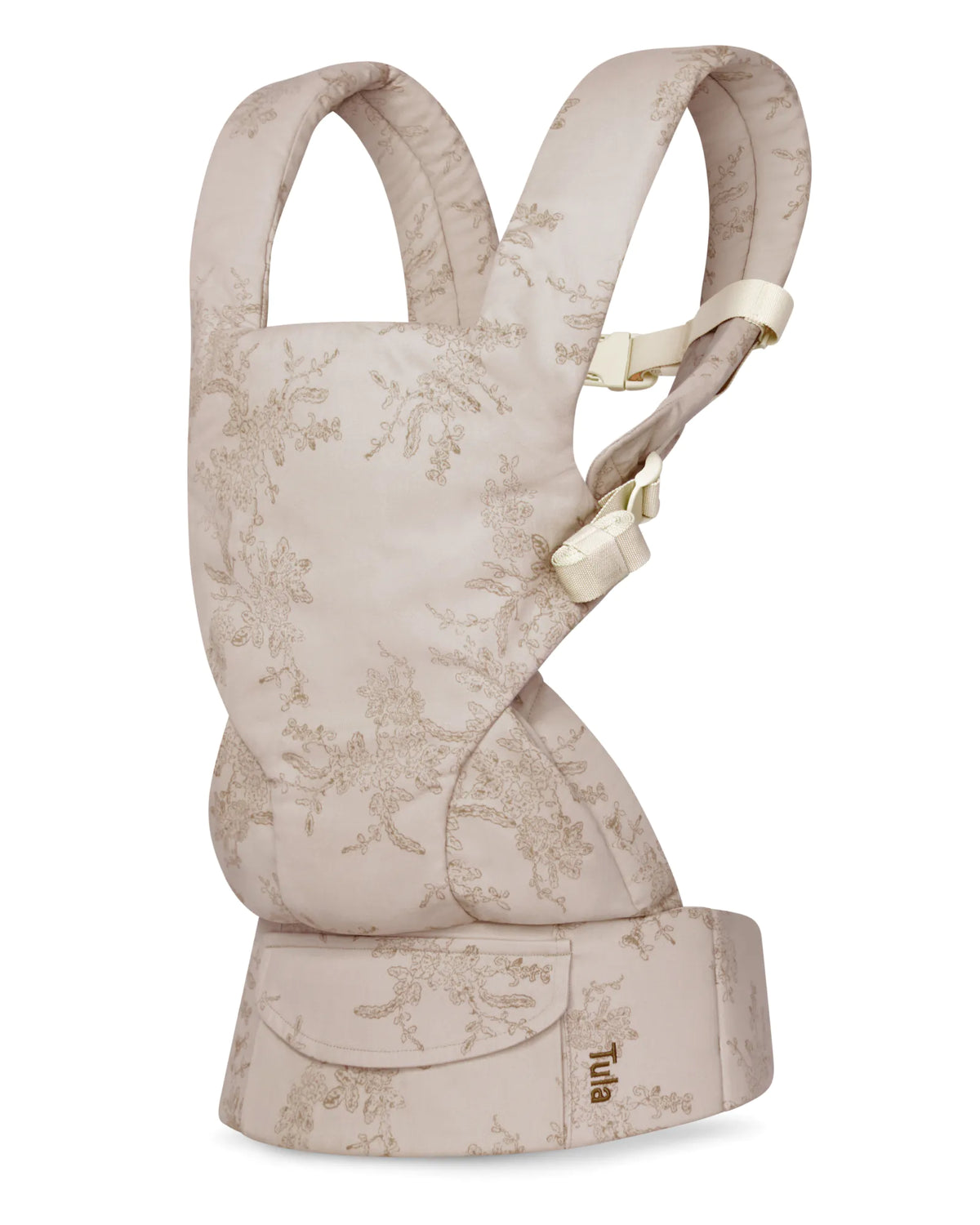 A Tula mesh baby carrier in a light beige fabric with a delicate floral toile pattern, featuring cream-colored buckle details and a matching front pouch.