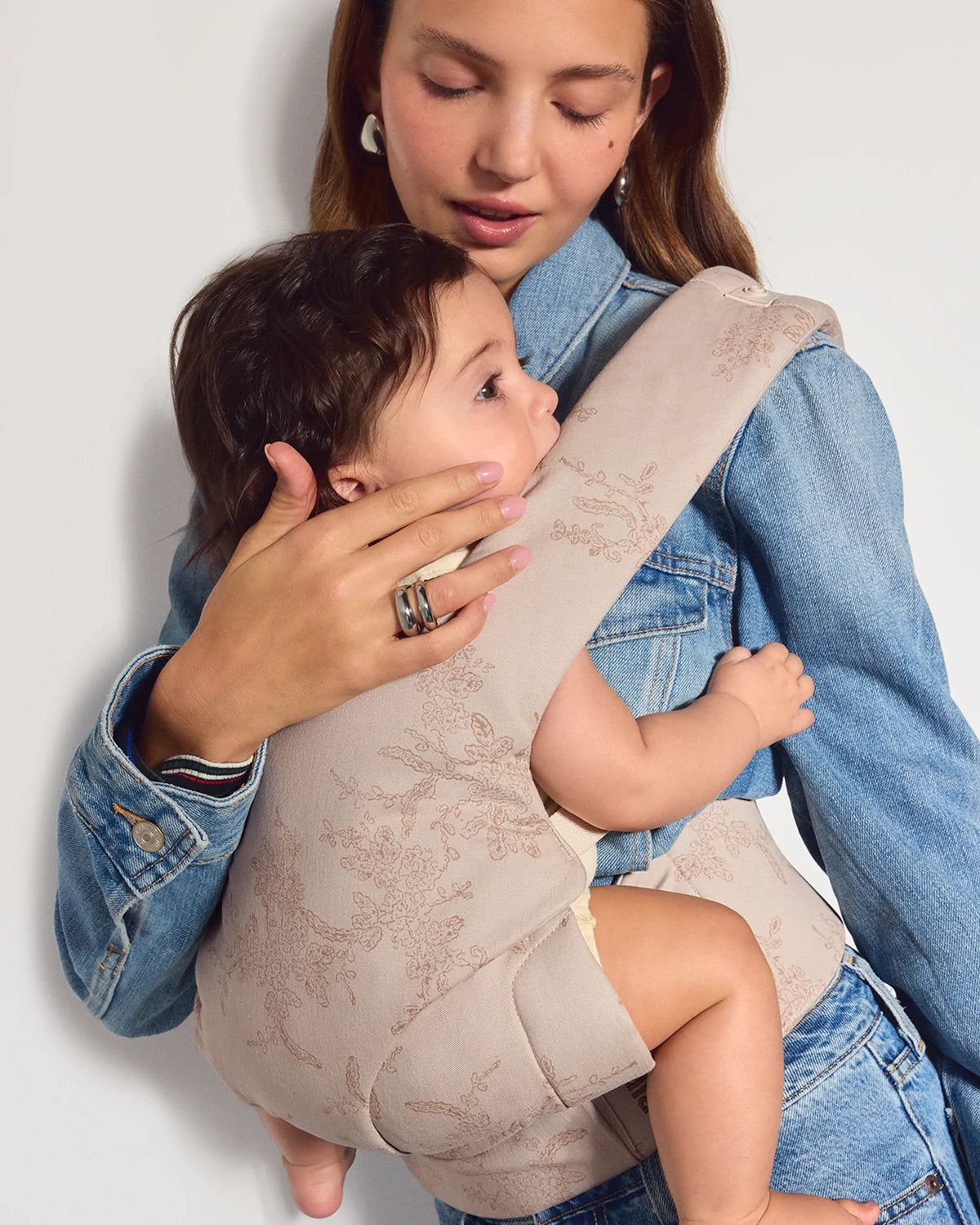 A woman wearing a light blue denim outfit with her baby wearing a beige knit explore baby carrier wrap against a plain light gray wall.