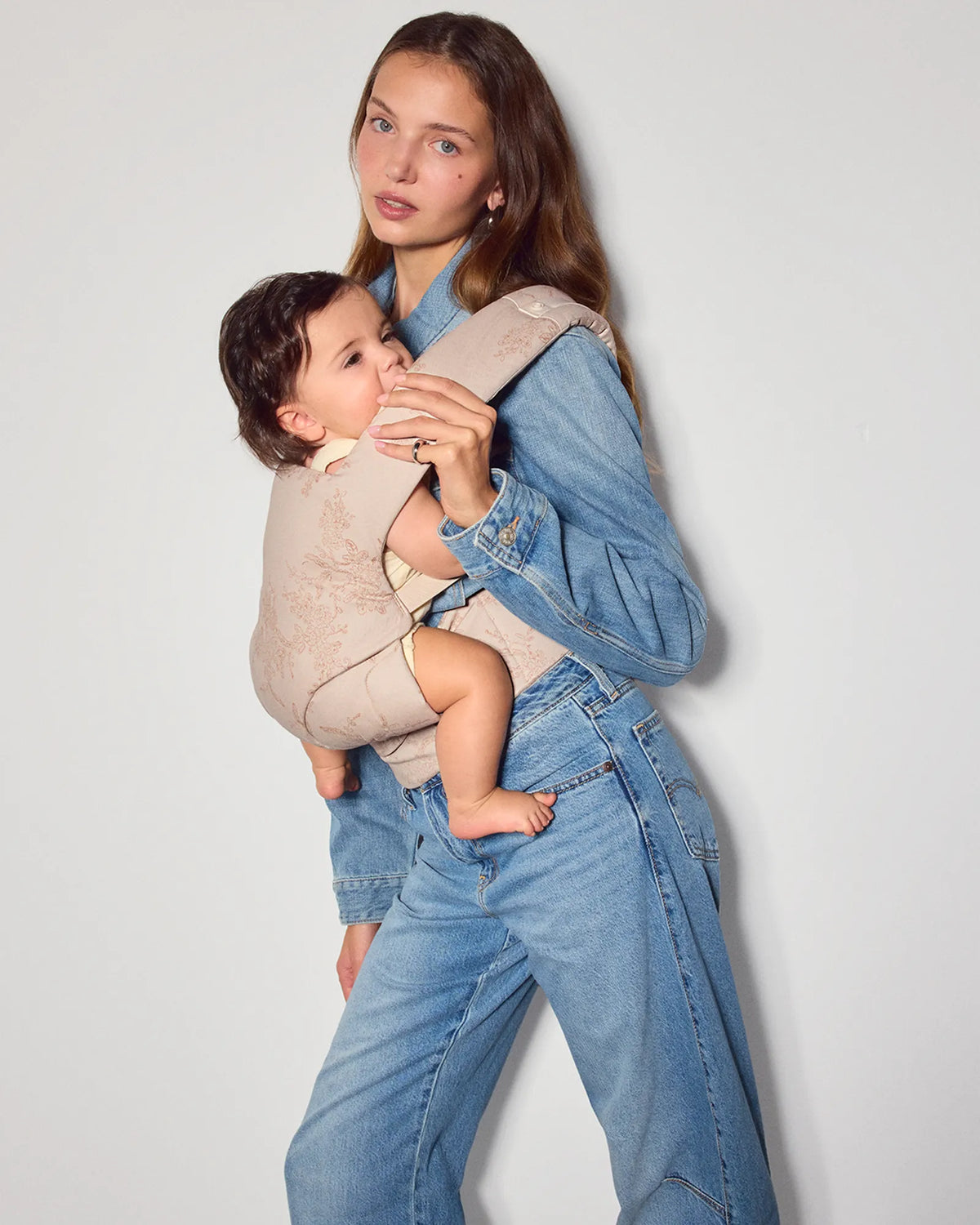 A woman wearing a light beige jacquard explore baby carrier with a subtle floral pattern, holding her baby inward facing. The woman is wearing a blue denim jacket and jeans, and the background is a clean white studio wall.
