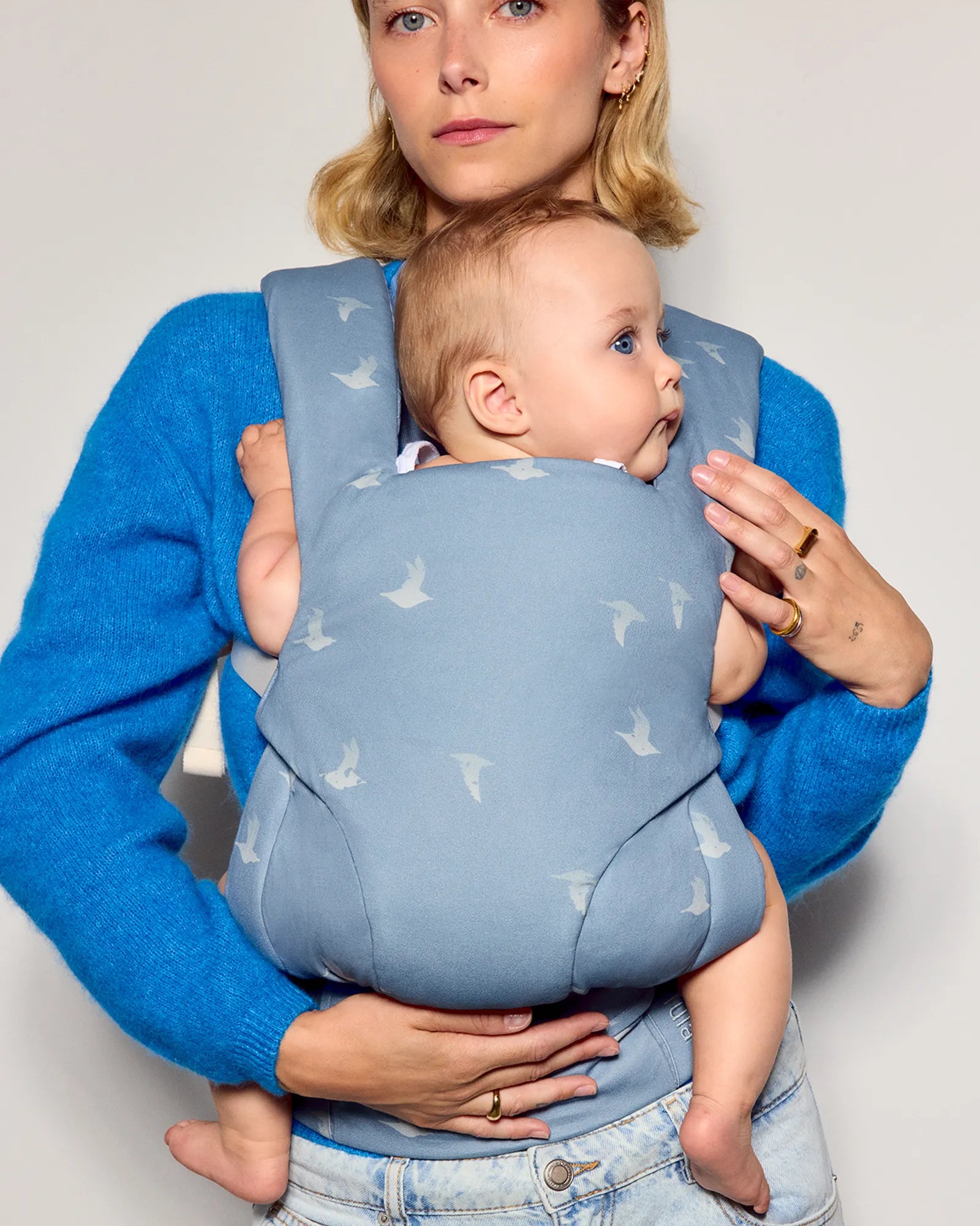 A woman wearing a bright blue sweater carries a baby in a front tula explore baby carrier. The carrier is a dusty light blue with a repeating white bird pattern.