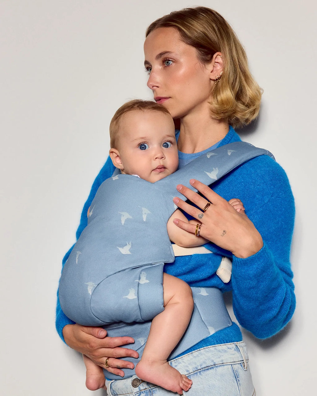 A woman wearing a bright blue sweater and light denim jeans carries a baby in a light blue tula explore baby carrier with a white bird pattern, set against a plain light gray studio background.