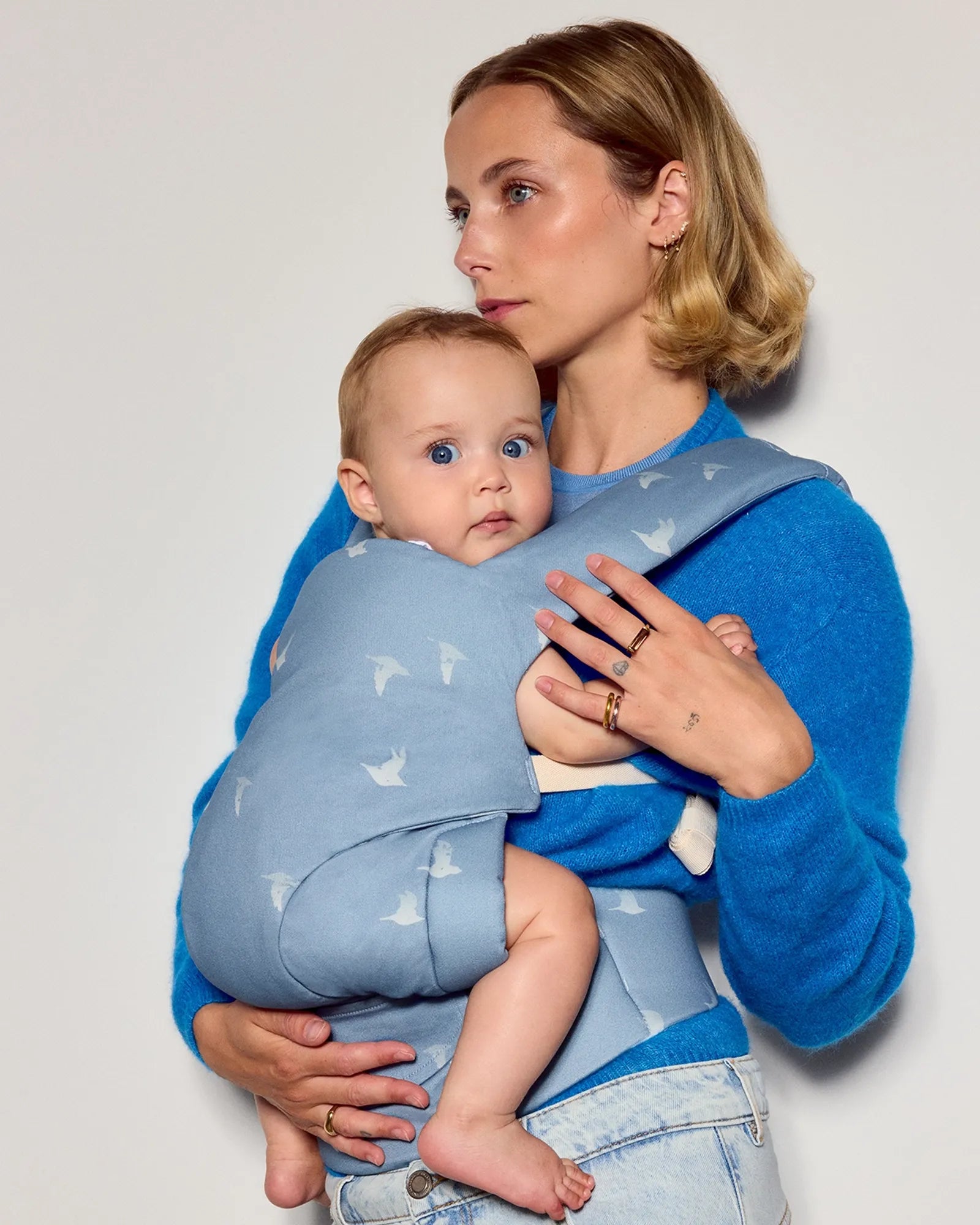 A woman wearing a bright blue sweater and light denim jeans carries a baby in a light blue tula explore baby carrier with a white bird pattern, set against a plain light gray studio background.