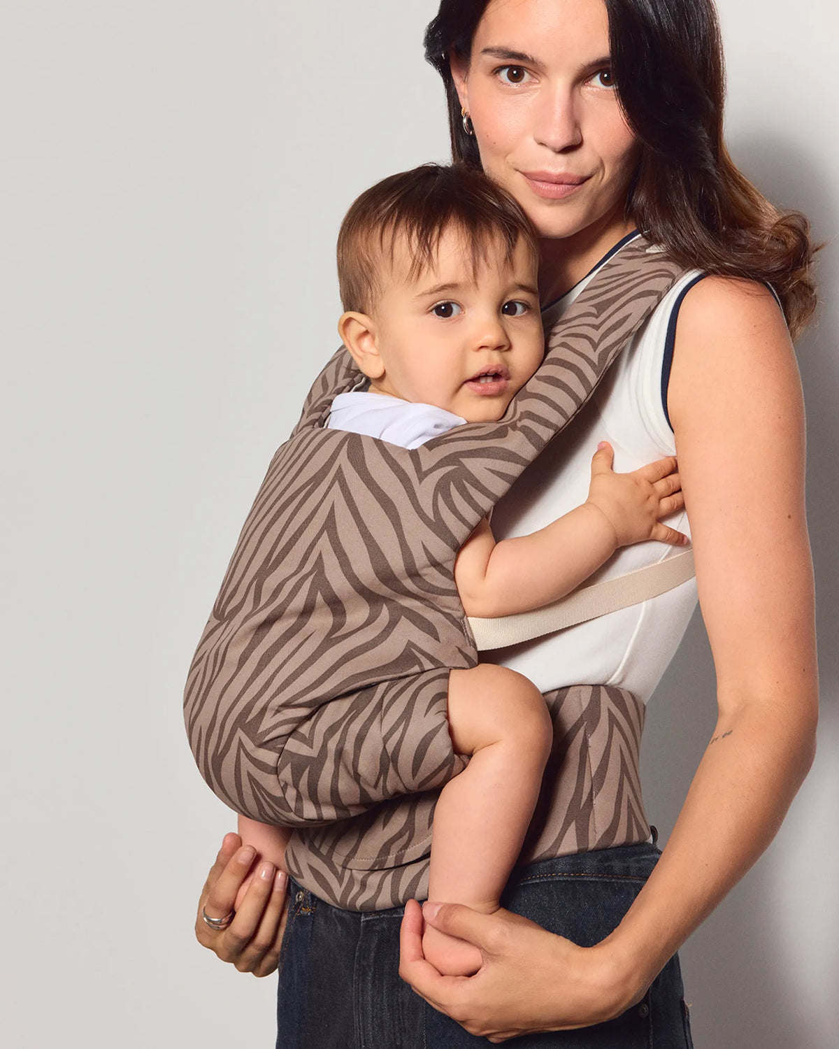 A woman wearing a brown and beige zebra-print baby carrier wrap with a baby, photographed against a plain light gray studio background.