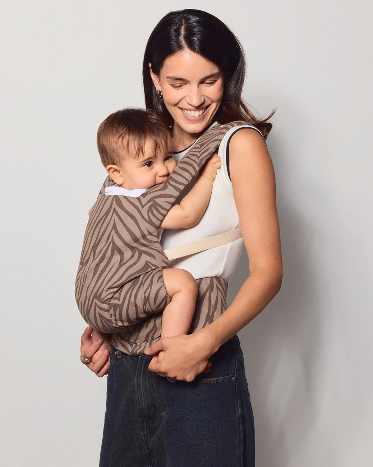 A woman wearing a dark denim skirt and white top holds a baby in a brown and beige zebra-print baby carrier against a light gray studio background.