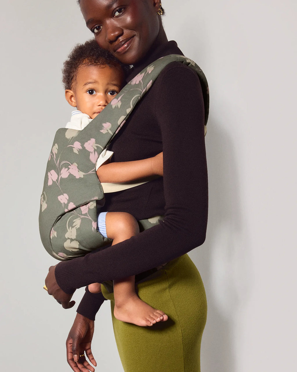 A woman wearing a black long-sleeve top and olive green pants carries a baby in a floral explore baby carrier. The carrier is sage green with a light pink and white floral pattern.