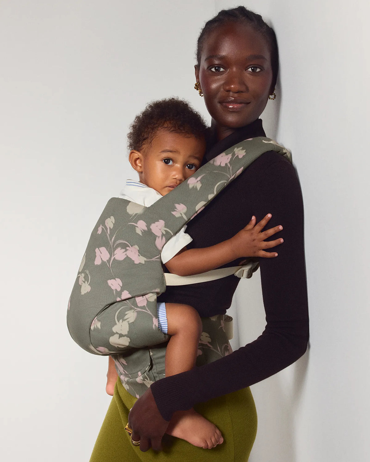 A woman wearing a dark long-sleeve top and olive green skirt carries a baby in a floral explore baby carrier. The carrier has a sage green base with a light pink and white floral pattern.