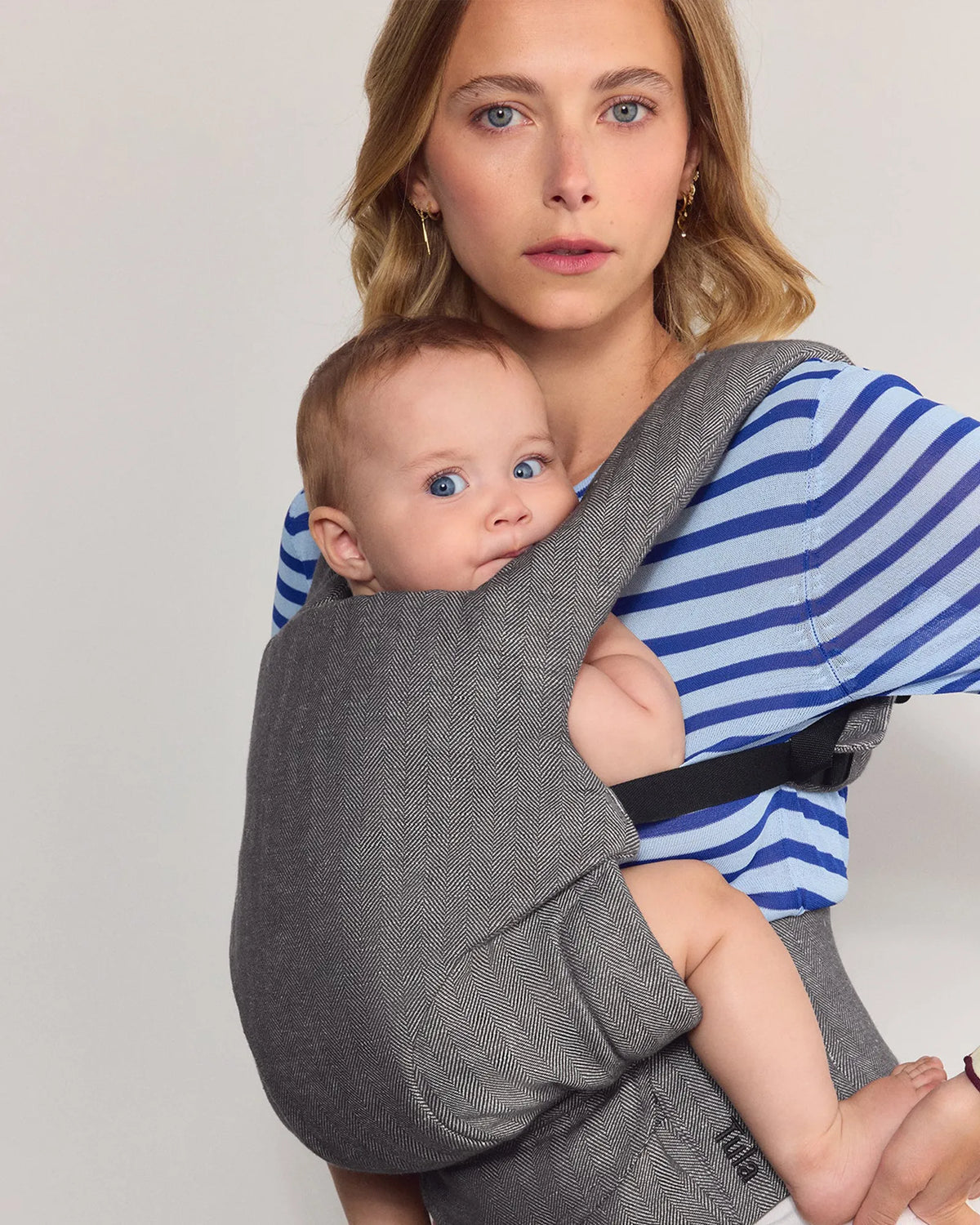 A woman wearing a blue and white striped long-sleeve shirt and white pants carries a baby in the tula explore baby carrier in ash. The photo is taken in a minimalist studio setting with a light gray wall and floor.