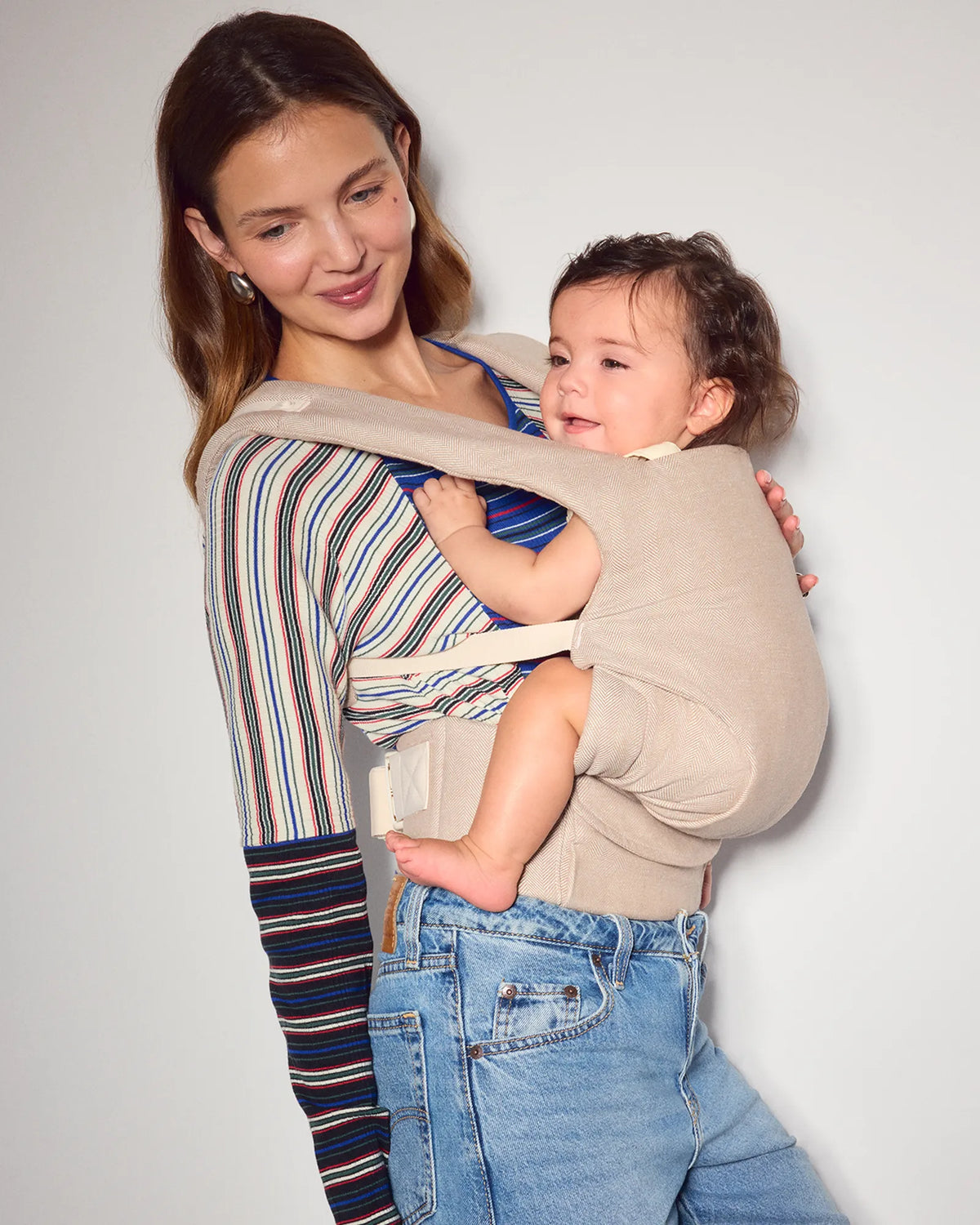A woman wearing the tula explore baby carrier in sand with a baby inside, paired with a striped long-sleeve top and blue jeans, against a plain light gray background.