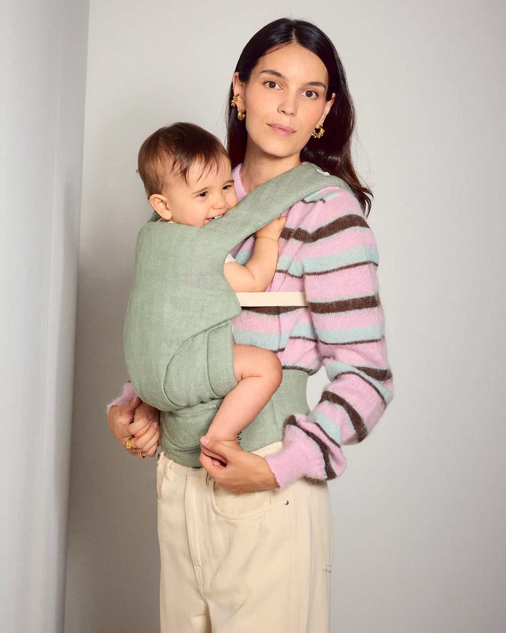 A woman wearing a pink, purple, and brown striped knit sweater carries a baby in a sage green tula explore baby carrier worn across her torso. 