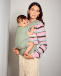 A woman wearing a pink, purple, and brown striped knit sweater carries a baby in a sage green tula explore baby carrier worn across her torso. 