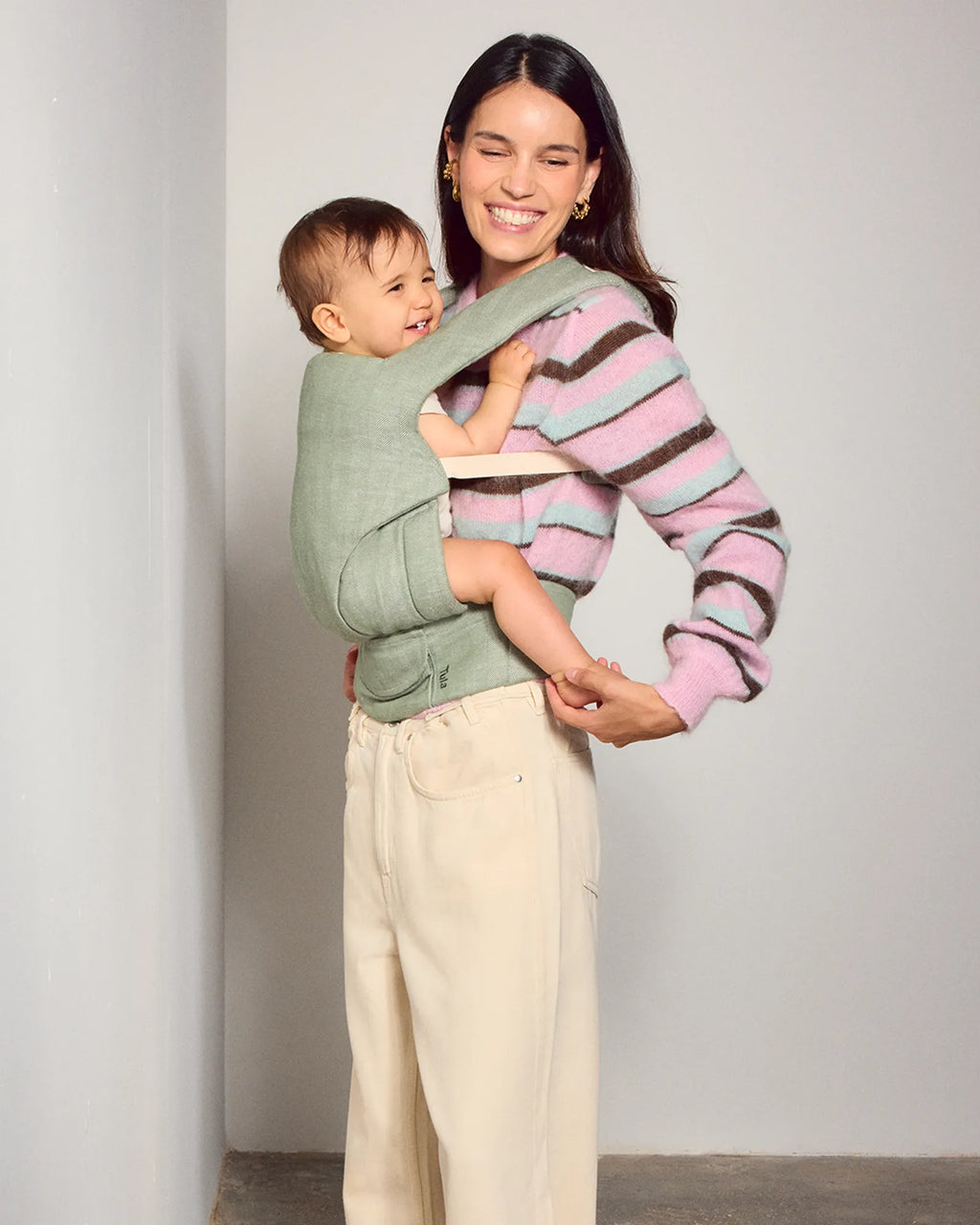A woman wearing a pink, purple, and brown striped knit sweater carries a baby in a sage green tula explore baby carrier worn across her torso. She is wearing cream-colored pants, standing against a neutral light gray wall.