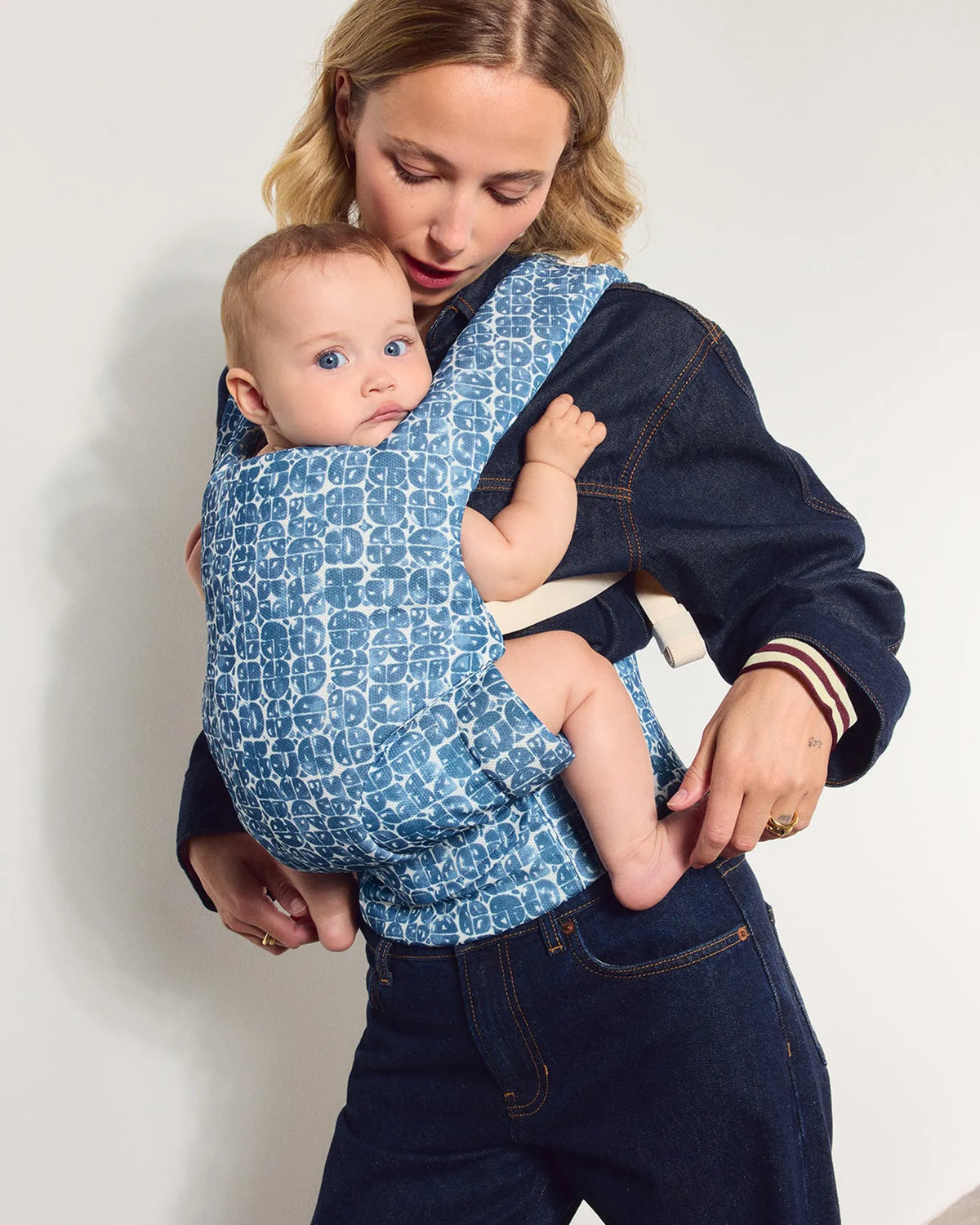 A woman wearing a dark denim outfit with a baby in a blue and white patterned mesh baby carrier, standing against a plain light gray wall.
