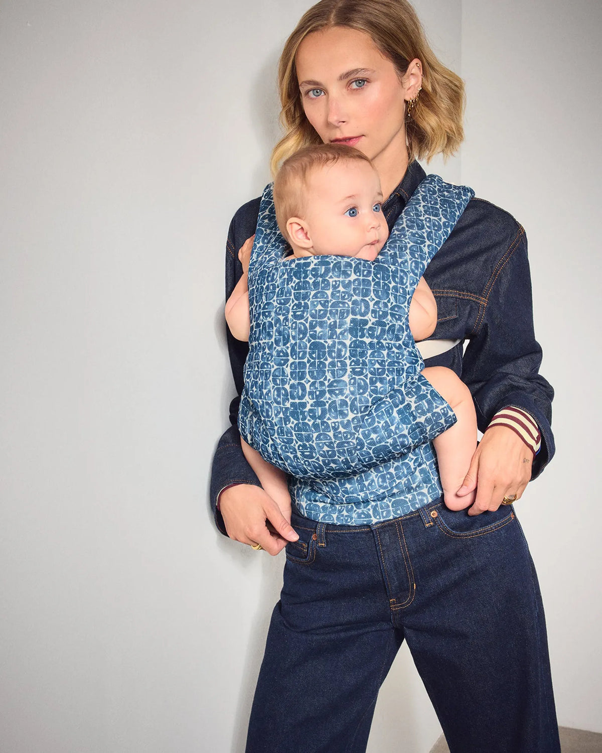 A woman wearing a blue patterned baby carrier with a baby, styled with dark denim clothing against a plain light gray wall.