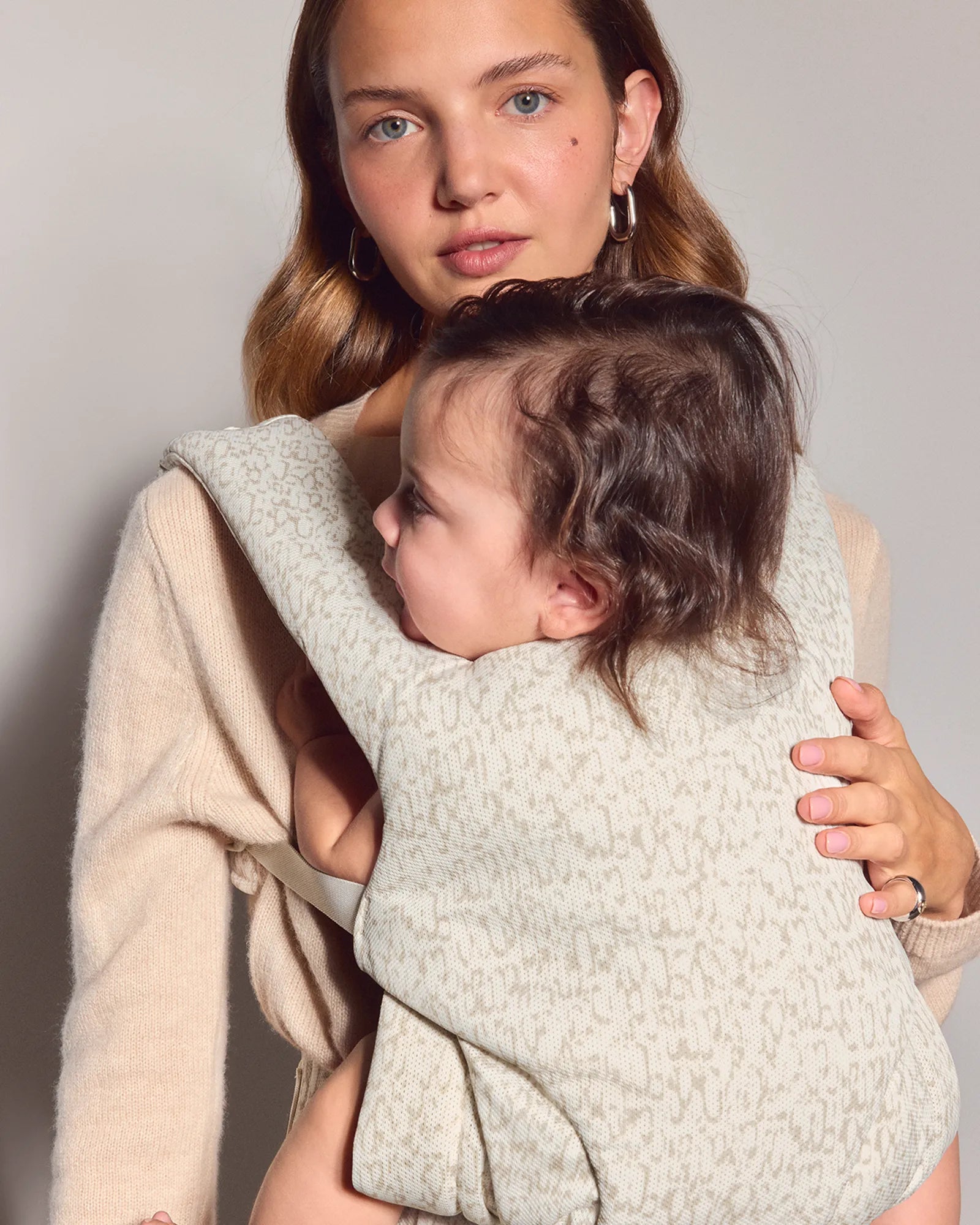 A woman wearing a beige cardigan carries a baby in a cream and beige patterned mesh explore baby carrier against a plain light gray studio background.