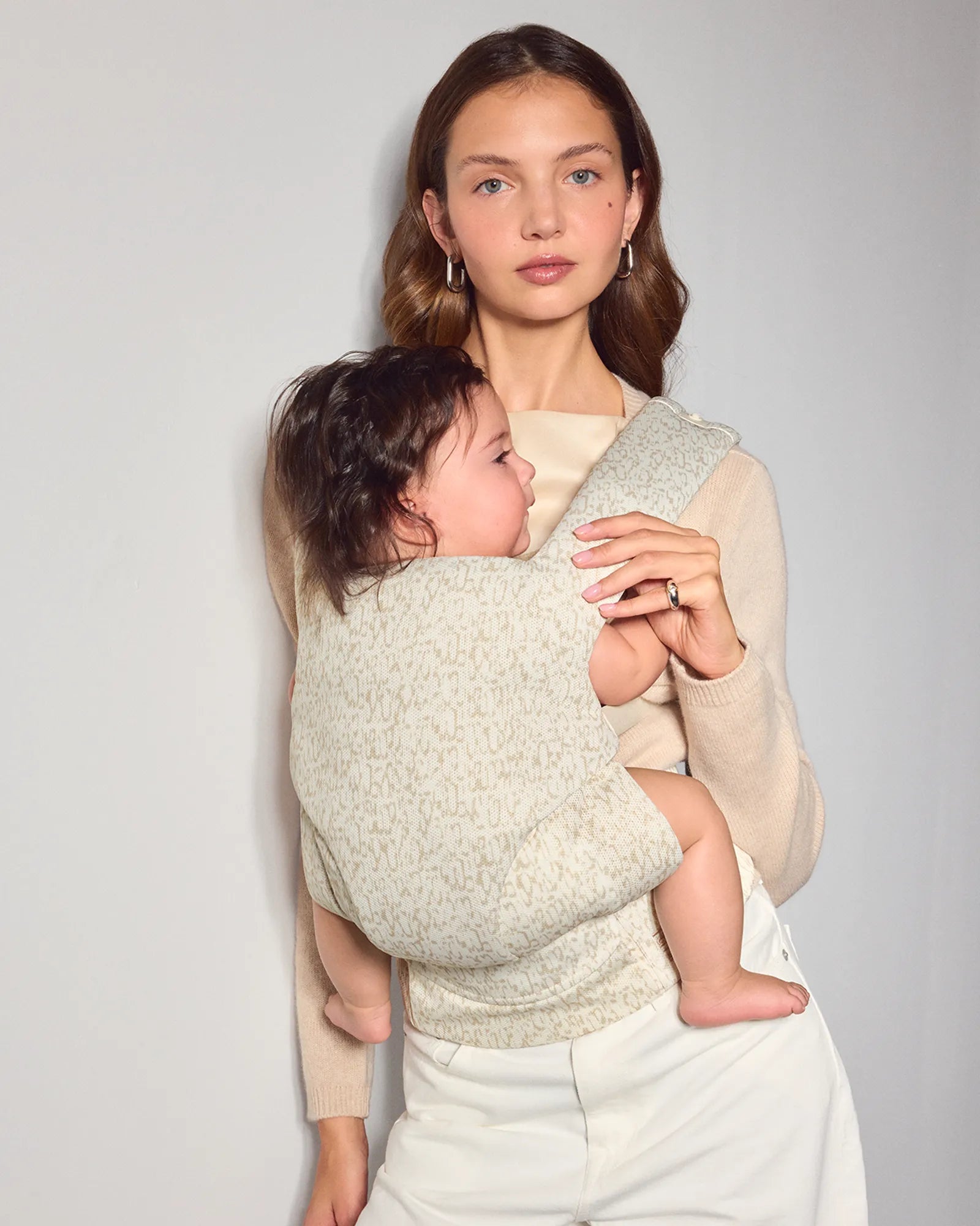 A woman wearing a light beige mesh baby carrier holds a baby wrapped in the same textured knit explore baby carrier against a plain light gray wall.