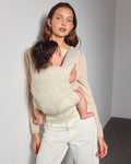 A woman wearing a sweater holds a baby in a cream and tan speckled mesh explore baby carrier. She is wearing light-colored trousers and a beige cardigan, standing against a neutral gray wall.