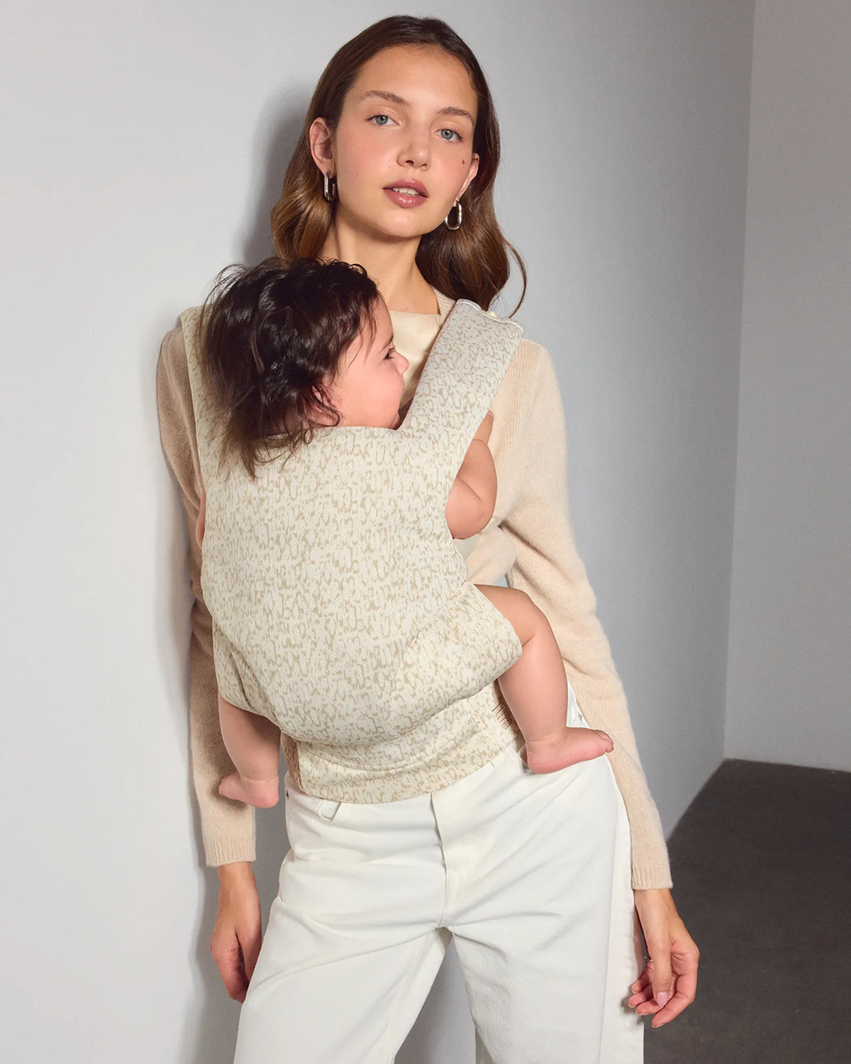 A woman wearing a sweater holds a baby in a cream and tan speckled mesh explore baby carrier. She is wearing light-colored trousers and a beige cardigan, standing against a neutral gray wall.