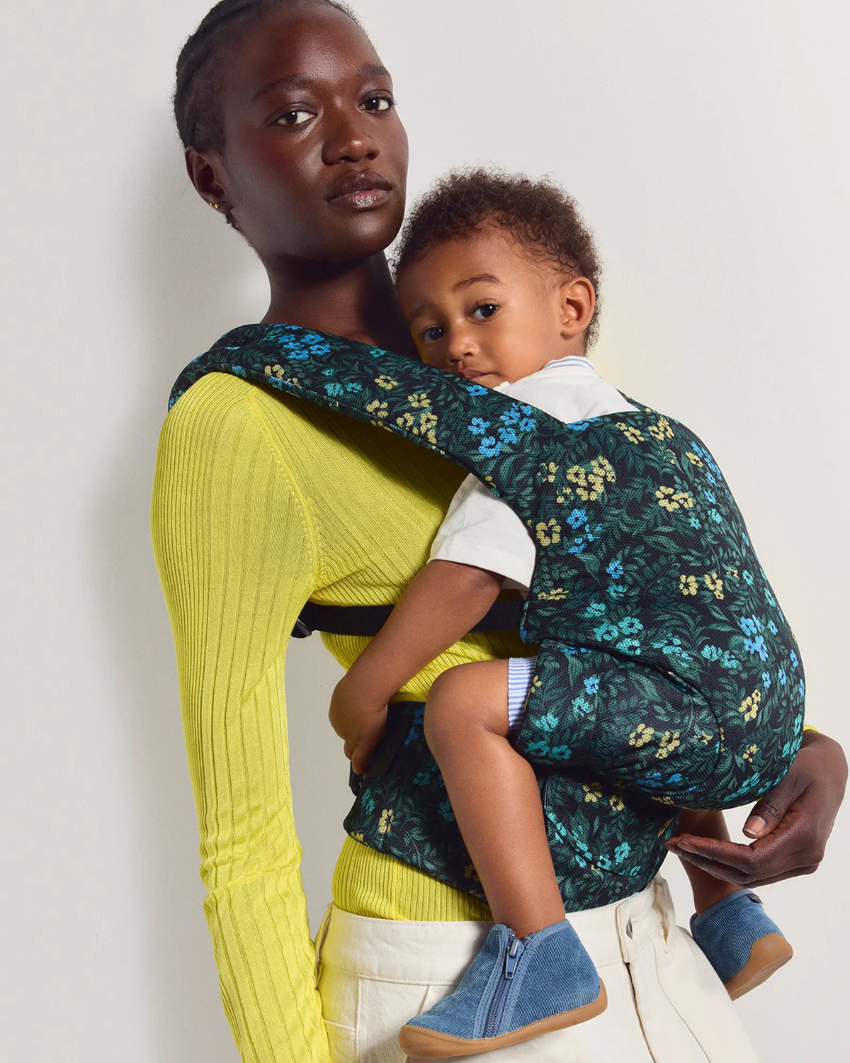 A woman wearing a bright yellow ribbed long-sleeve top and cream pants carries a baby in a structured front baby carrier. The carrier has a dark teal background with a floral pattern in yellow, blue, and green.