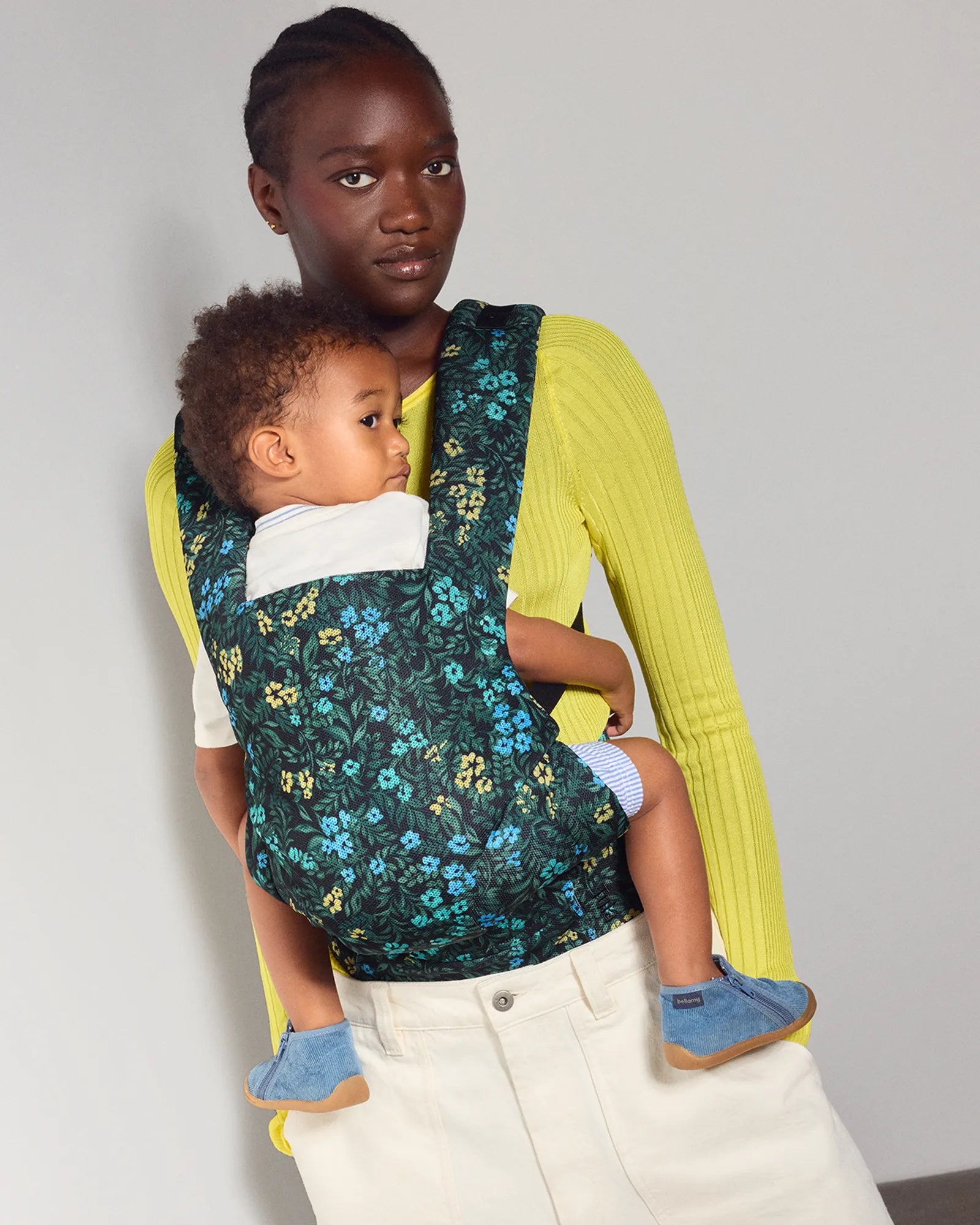 A woman wearing a bright yellow ribbed long-sleeve top and cream pants carries a baby in a dark green floral baby carrier. The baby is wearing light blue socks.
