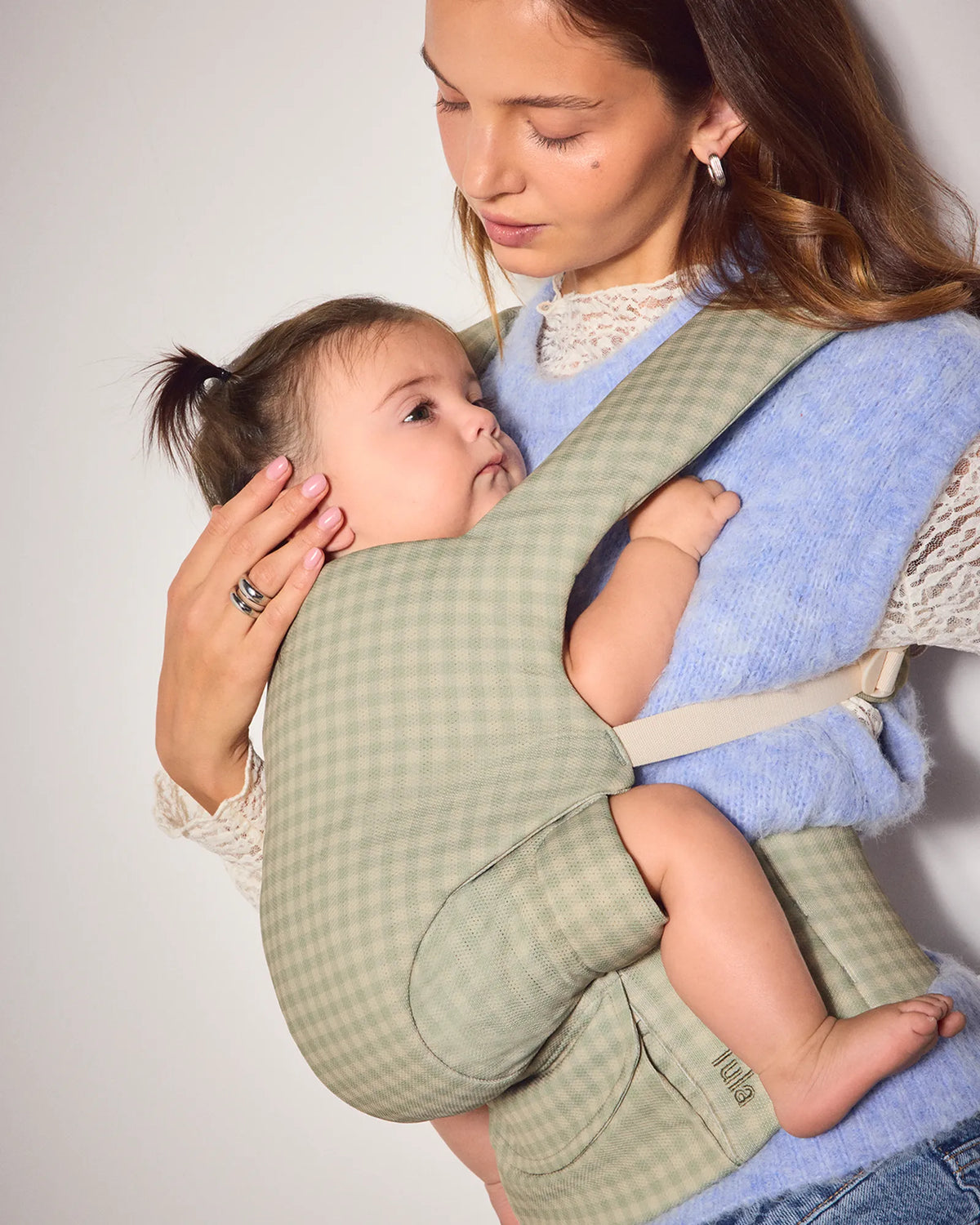 A woman wearing a light blue sweater and jeans holds a baby in a green and beige gingham check baby carrier against a plain light gray background.