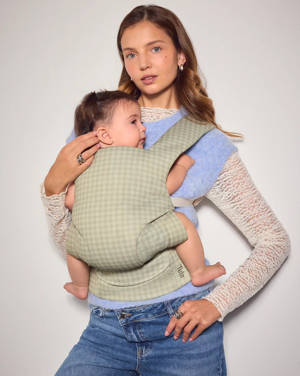 A woman wearing a cream lace-knit sweater and blue jeans holds a baby in a mesh baby carrier. The mesh baby carrier has a light sage green and white gingham pattern on the front panel and a soft light blue fabric lining.