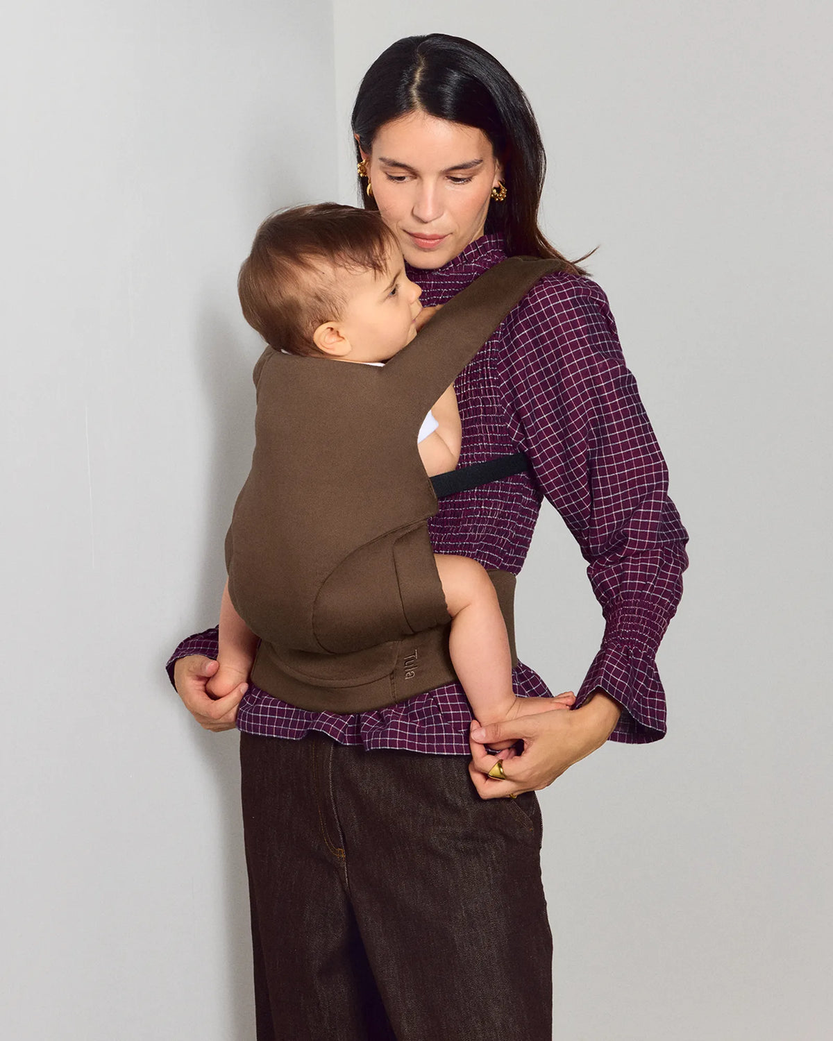 A woman wearing a cocoa tula explore baby carrier with a baby inside, styled with a purple plaid shirt and dark brown pants against a plain light gray background.