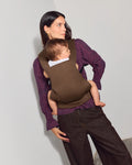 A woman wearing a cocoa tula explore baby carrier with a baby inside, styled with a purple plaid shirt and dark brown pants against a plain light gray background.