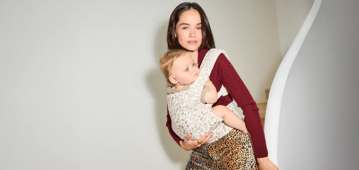 A woman carrying a baby in her tula explore baby carrier in snow cat print, wearing a burgundy long-sleeve top and a leopard print skirt against a minimalist indoor background.