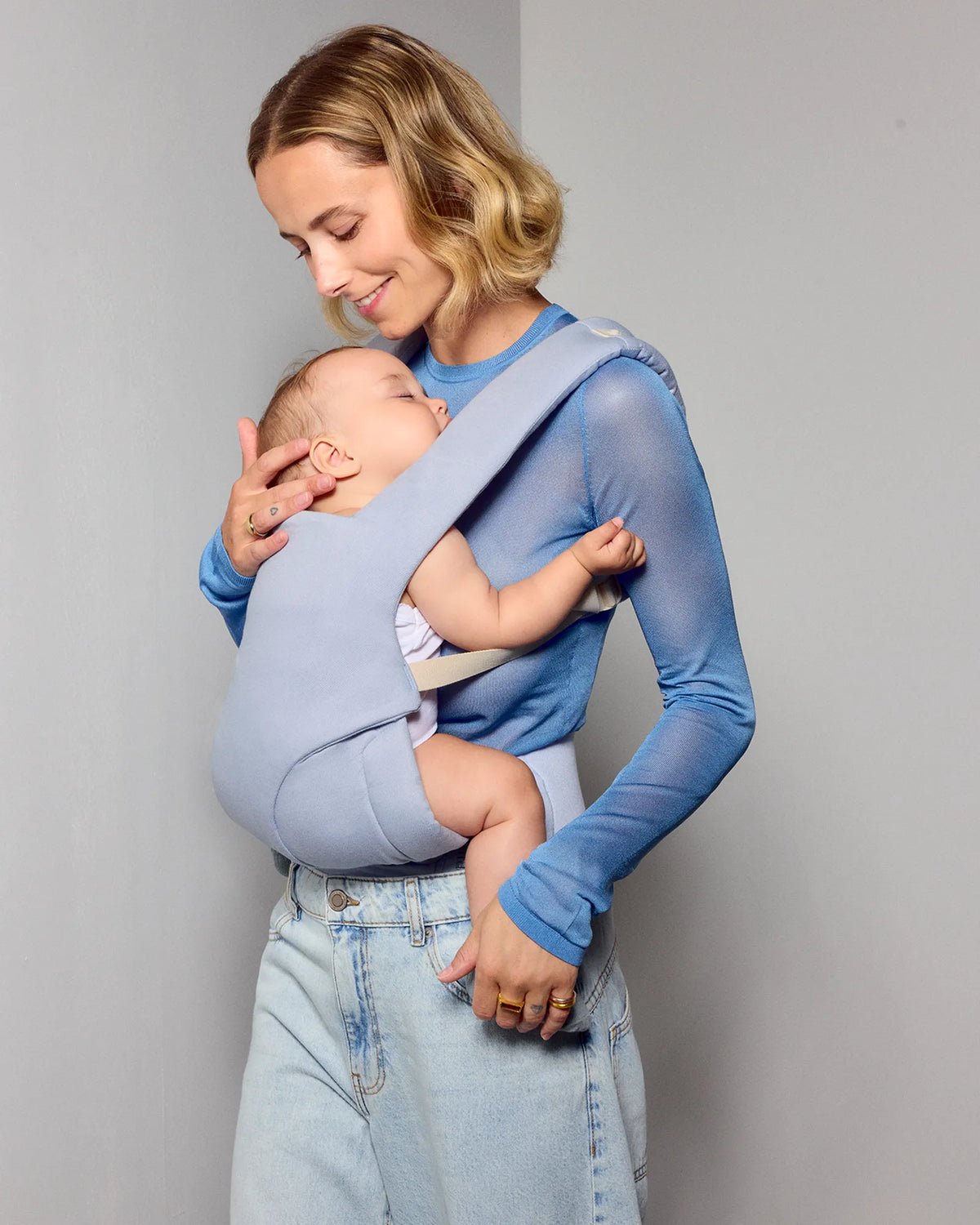 A woman wearing a light blue long-sleeve shirt and light-wash jeans holds a baby in a pale blue wrap tula explore baby carrier against a plain gray studio background.