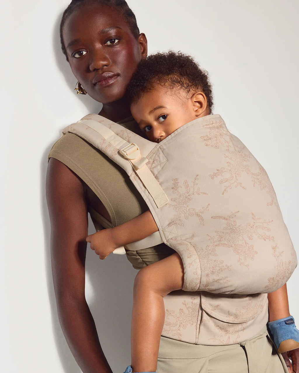 A woman wearing a tula original baby carrier in chantilly botanica with a child on her back, photographed against a plain light background.