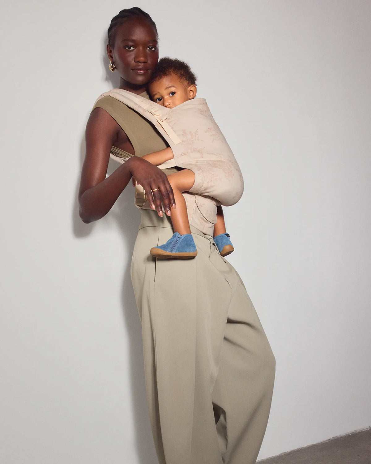A woman wearing a beige baby carrier wrap and wide-leg trousers holds a baby wrapped in a matching beige wrap. The woman is standing against a plain light gray wall on a concrete floor.