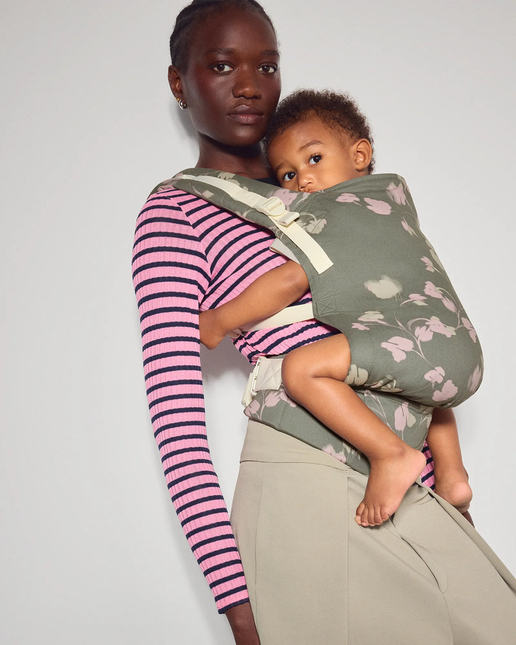 A woman wearing a pink and purple striped long-sleeve shirt and khaki pants carries her baby in the tula original baby carrier in Petal. The carrier is a muted sage green with a light pink and white floral pattern.