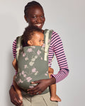 A woman wearing a pink and purple striped long-sleeve shirt and khaki pants carries her baby in the tula original baby carrier in Petal. The carrier is a muted sage green with a light pink and white floral pattern.