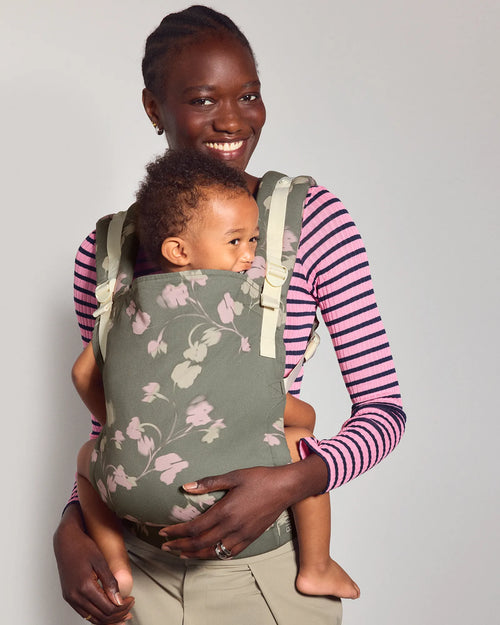 A woman wearing a pink and purple striped long-sleeve shirt and khaki pants carries her baby in the tula original baby carrier in Petal. The carrier is a muted sage green with a light pink and white floral pattern.