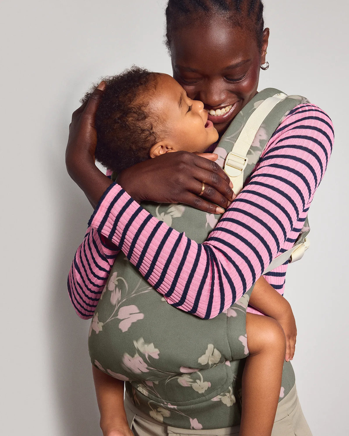 A woman wearing a pink and navy striped long-sleeve top and a sage green tula original baby carrier with a floral print, holding and kissing her baby against a plain light gray background.