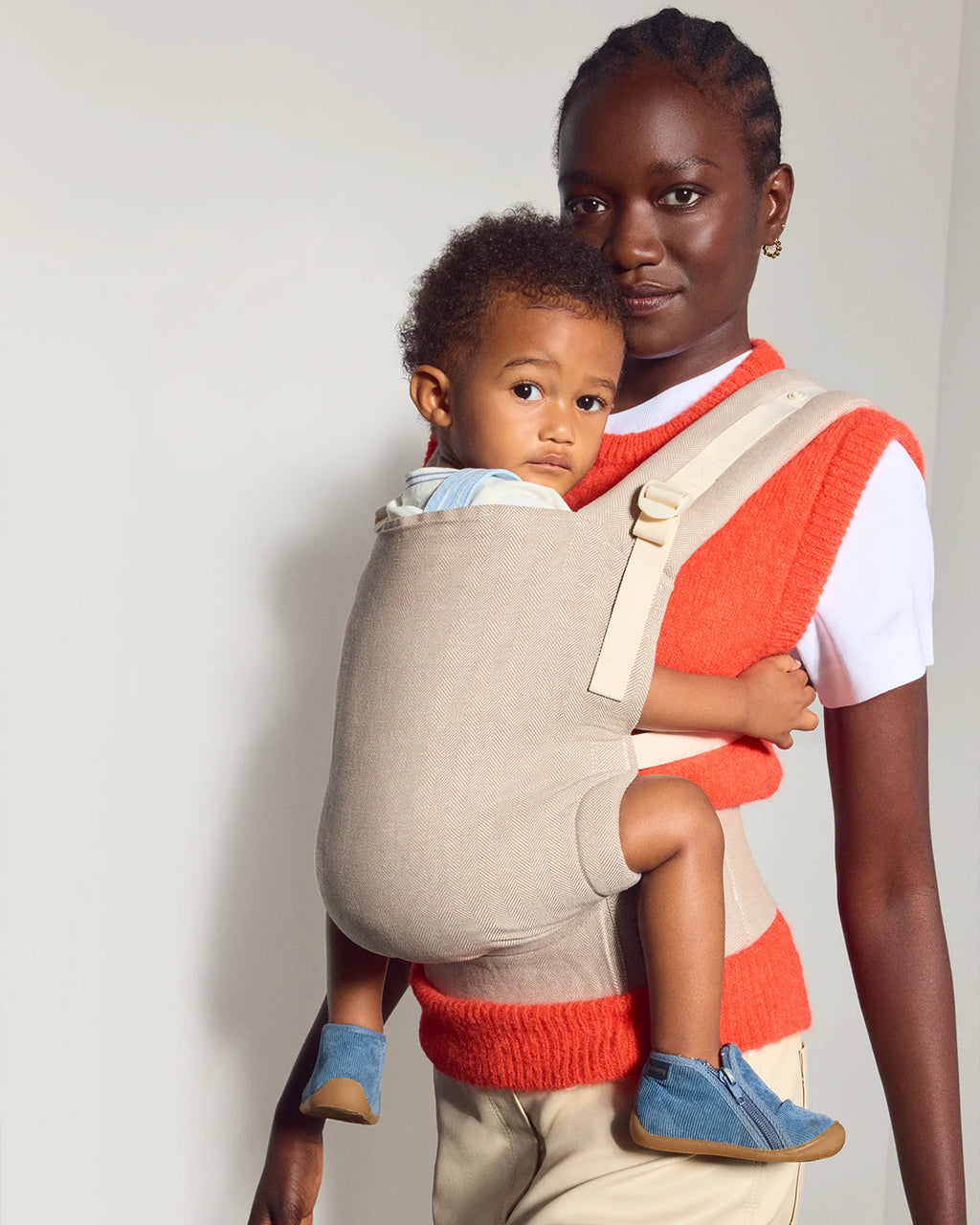A woman wearing a bright orange knit sweater vest over a white t-shirt and beige pants carries a young child in a the tula original baby carrier. The woman is standing against a plain light gray wall.