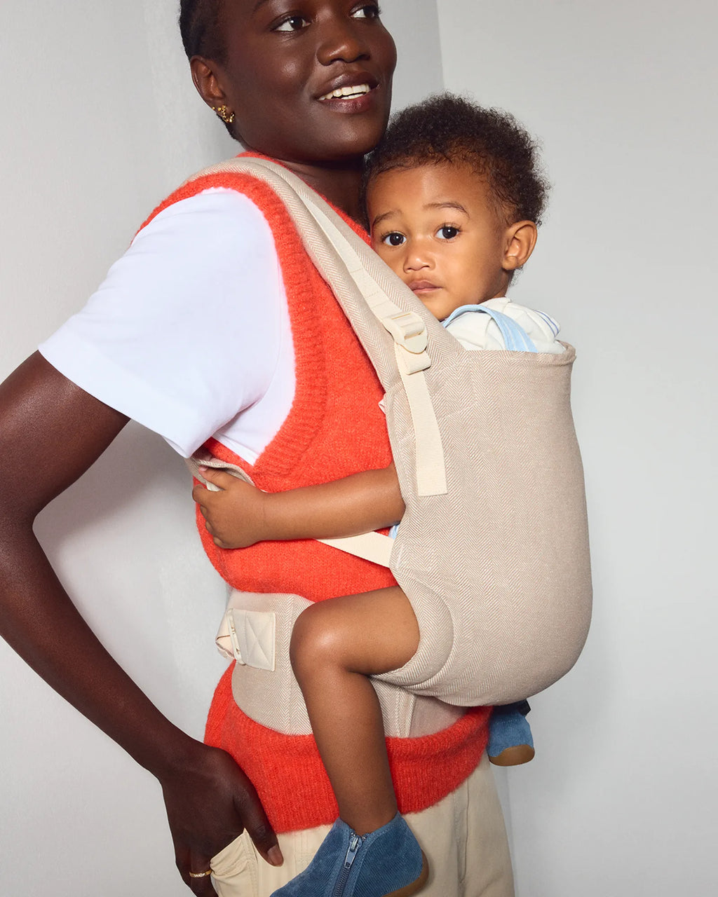 A woman wearing a bright coral-red knit vest and white T-shirt carries a baby in a beige front original baby carrier. The baby is wearing a light blue and white striped shirt and blue shoes.