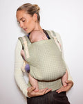 A woman wearing a light sage green and cream tula original baby carrier with a baby inside, shown against a plain white background.
