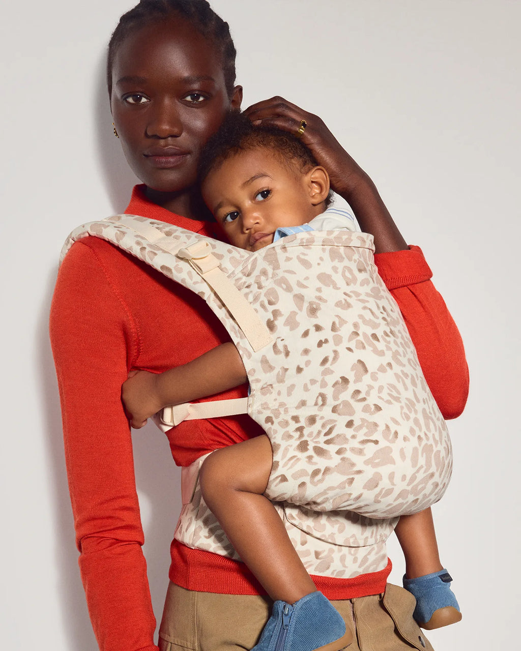 A woman wearing a bright red long-sleeve top and khaki pants holds a baby in a cream-colored baby carrier with a brown animal print pattern. The baby is wearing blue shoes.