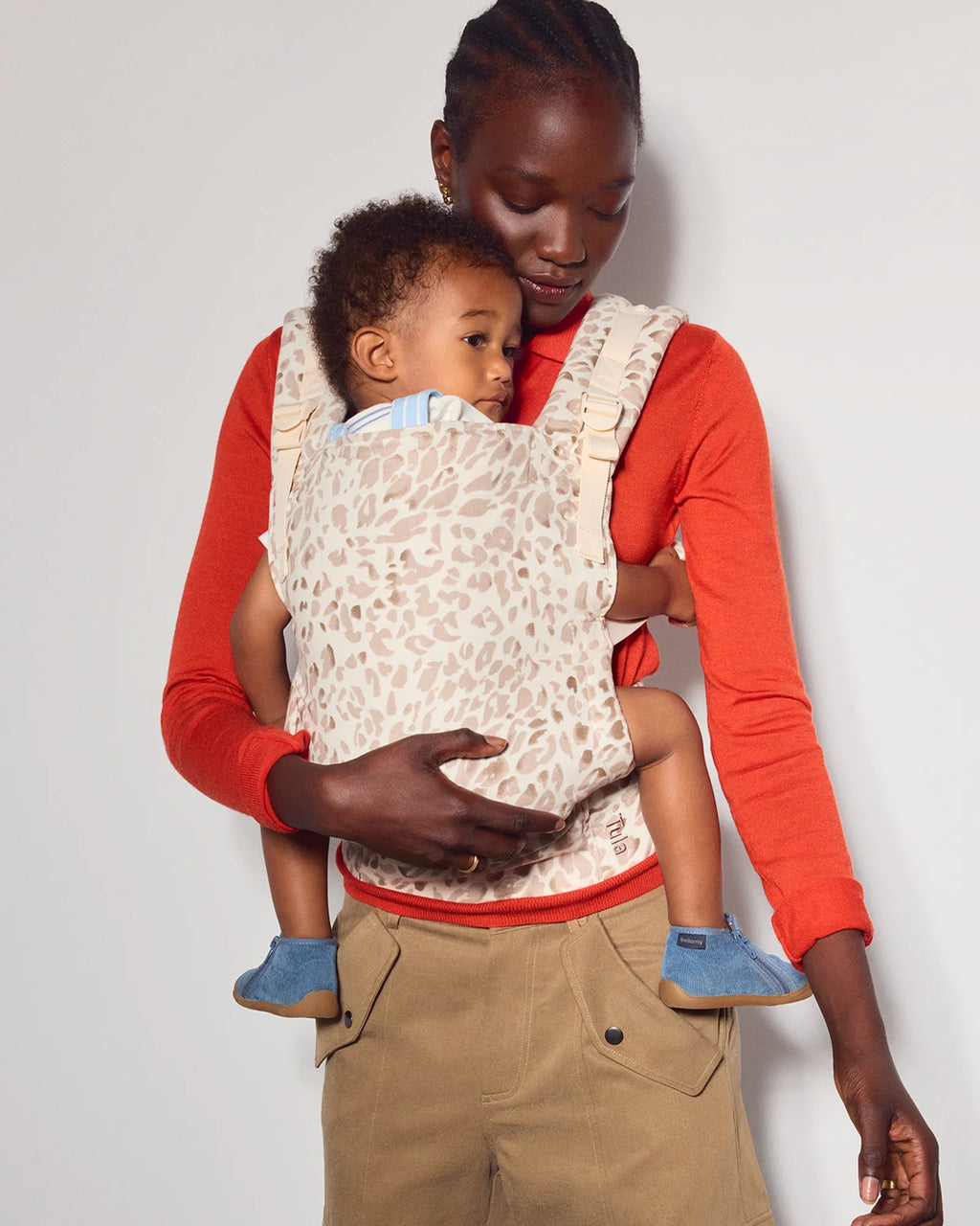 A woman wearing a red long-sleeve shirt and tan cargo pants carries a baby in a cream leopard-print baby carrier wrap against a plain light gray background.
