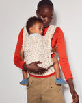 A woman wearing a red long-sleeve shirt and tan cargo pants carries a baby in a cream leopard-print baby carrier wrap against a plain light gray background.