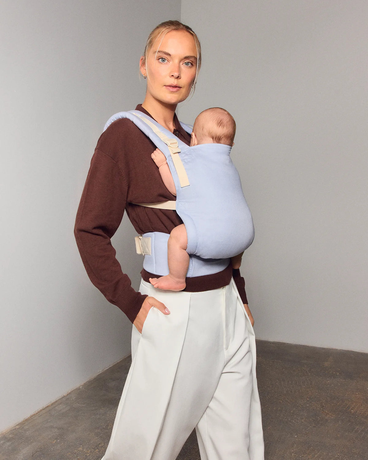 A woman wearing a brown long-sleeve top and light gray pants carries a baby in a light blue front tula original baby carrier. The baby is wrapped in a pale blue fabric and rests against the woman's torso.