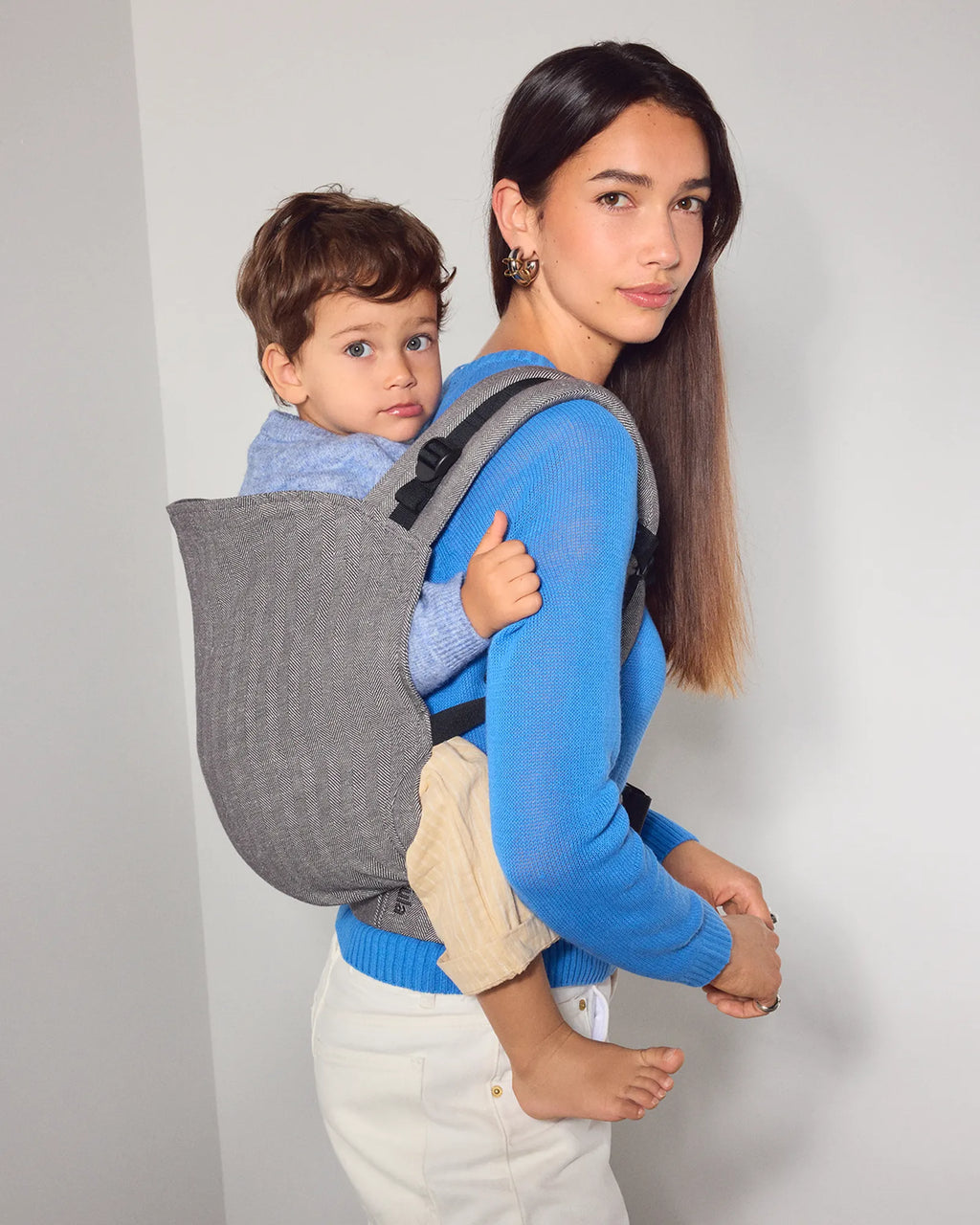 A woman wearing a bright blue sweater and white pants carries a child in a gray fabric toddler carrier on her back, standing against a plain light gray wall.