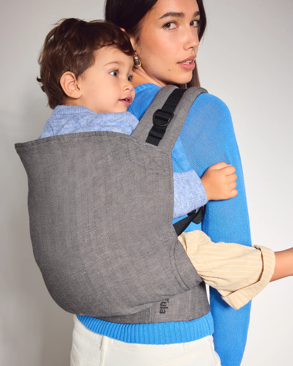 A woman wearing a bright blue sweater carries a child in a gray woven baby carrier on her back. The toddler carrier has black adjustable shoulder straps and a black buckle, and the brand name 'mima' is visible near the bottom corner.