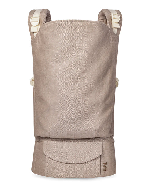 A Tula toddler carrier in a light beige linen fabric, featuring cream-colored shoulder straps and a front pocket with the Tula brand name printed in black.