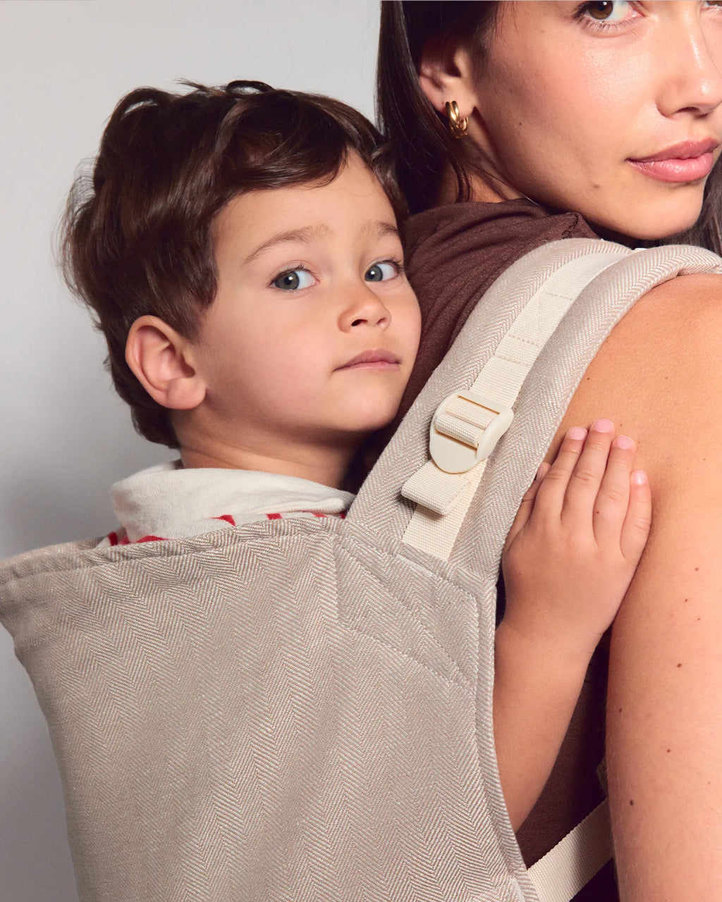 A close-up of a woman wearing a beige toddler carrier with a young child inside, shown against a plain light gray background.
